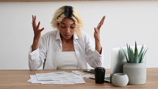 Broke,Frustrated african american girl holding her head sitting in front of pile of bills and invoices.Expenses,cost of living,debt concept. 