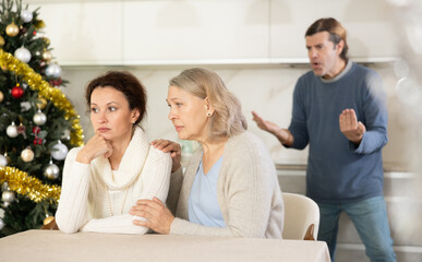 Man on christmas eve quarrels with his wife in kitchen, standing behind her, and elderly mother hugs and calms her daughter