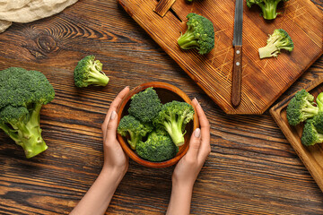 Female hands with bowl of fresh broccoli cabbages on wooden background - obrazy, fototapety, plakaty