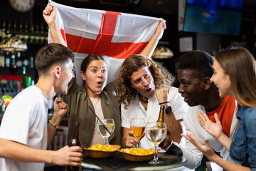 Group of cheerful football fans rooting for England team together in sports bar, waving national...