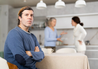 Frustrated man sitting in kitchen during quarrel between his wife and elderly mother. Complicated...