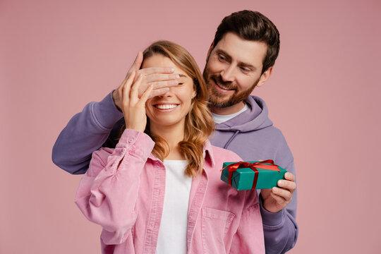 Handsome smiling man holding gift box, closed eyes of young beautiful woman. Portrait of happy romantic couple celebration birthday isolated on pink background. Dating, love, Valentines day