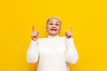 old cheerful grandmother with short hair smiling and pointing with her hands up on a yellow isolated background, elderly pensioner showing and advertising copy space
