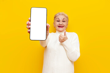old funny grandmother in a white sweater shows a blank smartphone screen and like on a yellow...