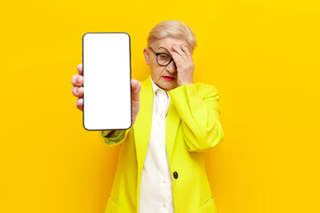 old unhappy grandmother in business clothes and blazer shows blank screen of smartphone and loses...