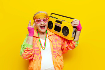old funny grandmother in a youth costume dances with a tape recorder and listens to music on yellow...