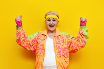 old funny grandmother in a youth tracksuit rejoices in victory with raised hands on a yellow isolated background, elderly woman pensioner in a stylish outfit celebrates and wins