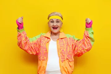 Cercles muraux Vielles portes old funny grandmother in a youth tracksuit rejoices in victory with raised hands on a yellow isolated background, elderly woman pensioner in a stylish outfit celebrates and wins