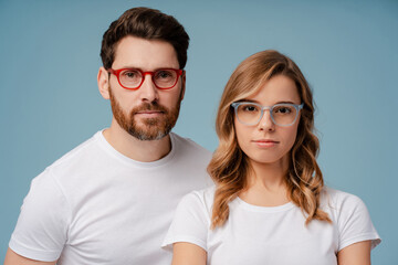 Closeup portrait of attractive serious  man and woman wearing white t shirt and stylish eyeglasses isolated on blue background. Confident fashion models looking at camera posing for pictures in studio