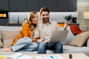 Smiling young couple using laptop computer holding credit card shopping online sitting on sofa at home. Attractive man and woman, freelancer working online receive payment - obrazy, fototapety, plakaty