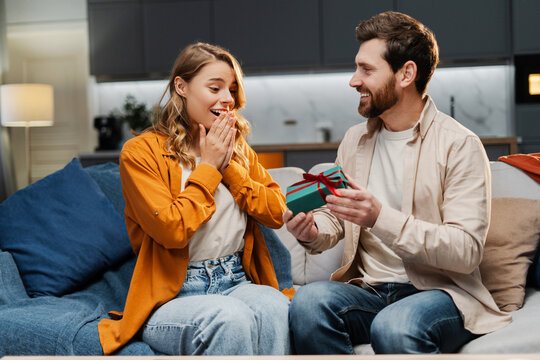 Bearded handsome man giving gift box to beautiful excited woman at home. Happy romantic couple celebration birthday. Dating, love, Valentines day concept
