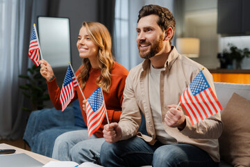 Smiling American couple of friends holding American flags watching tv celebration Independence day...