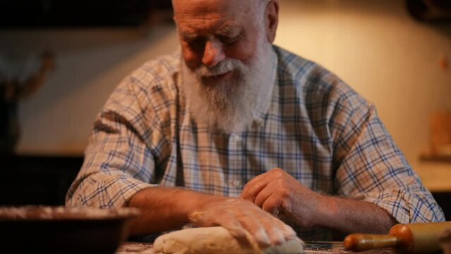 Close-up. An elderly bald man with a beard in a shirt sprinkles flour and rubs with his hand an oblong piece of dough lying on a wooden table. The man smiles and talks to the dough