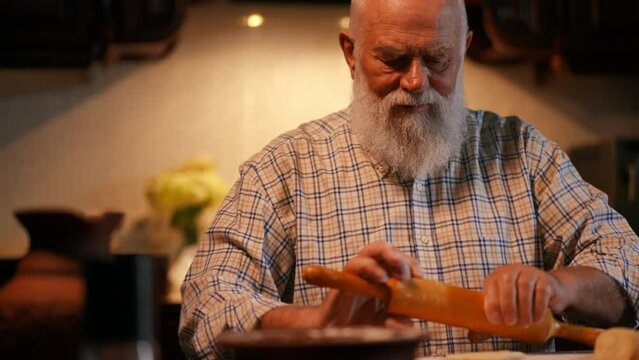 Close-up. An elderly bearded man in a shirt rolls out dough with a rolling pin on a wooden board. A man sprinkles flour on the rolled out dough. Making dumplings in the home kitchen