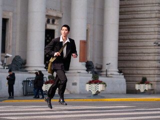 Obraz premium Portrait of handsome Chinese young man with black short hair wearing black blazer running across street with modern city building background in sunny winter day, male fashion, cool Asian young man.
