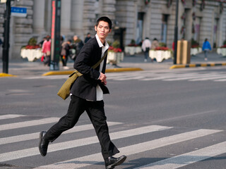 Portrait of handsome Chinese young man with black short hair wearing black blazer running across street with modern city building background in sunny winter day, male fashion, cool Asian young man.