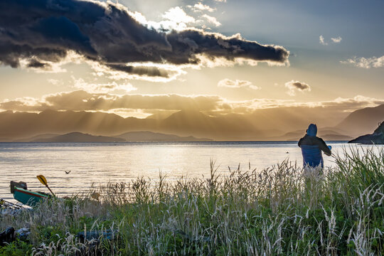 sunset in the chonos archipelago: a sea kayaker on a beach  stands in silhouette looking out over a wide channel to a dramatic sunset over mountains in the distance room for text
