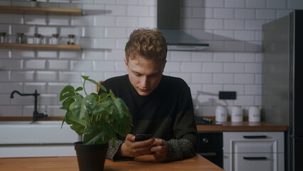Young handsome man sitting in the modern kitchen, looking at his smartphone and checks his messages	
