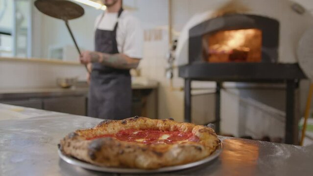 Chef serving hot pizza with bubbly crust and salami from the peel on a steel plate on table in pizzeria kitchen. Close-up view