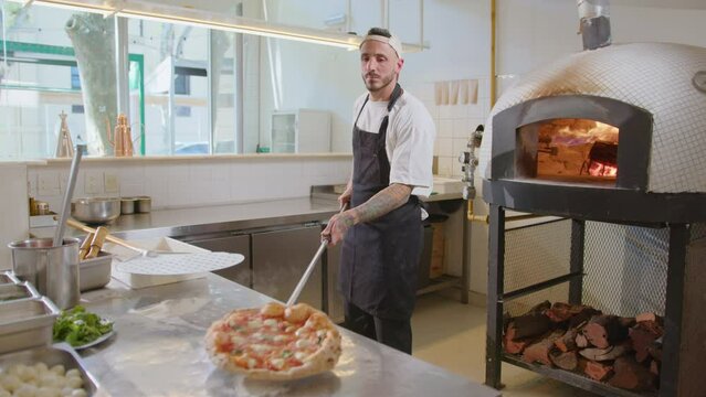 Young chef in apron taking hot baked pizza from wood-fired oven and putting it on the table in pizzeria kitchen
