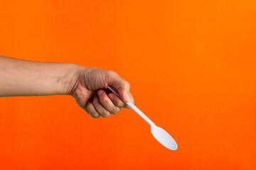 White plastic spoon in hand on orange background - obrazy, fototapety, plakaty