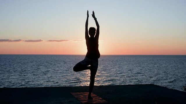 Woman practicing yoga standing in tree pose asana on sea beach at sunset. Silhouette of female on sport mat on nature nightfall. Sports, yoga practice, stretch, wellness, healthy lifestyle concept.