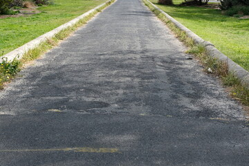 a weathered asphalt path with signs of wear and patchwork, and overgrown grass on either side of...