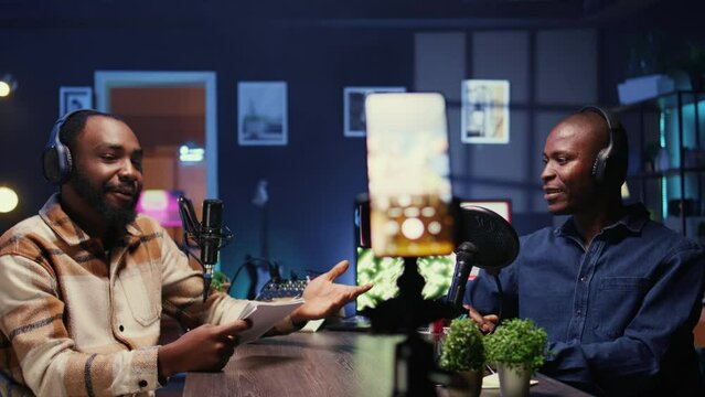 Cohosts streaming podcast using smartphone on tripod during live debate on vlog channel, handheld camera shot. Men meeting in studio to film show with mobile phone and talk politics