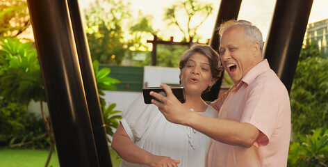 Indian Asian Hindu happy old older gen x male female couple standing hold mobile use phone look cam...
