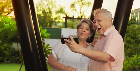 Indian Asian Hindu happy old older gen x male female couple standing hold mobile use phone look cam take funny faces selfie outdoor garden aged elder man woman lady talk gossip enjoy fun joy home park