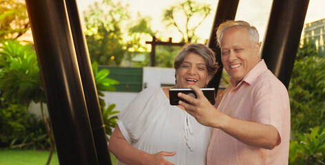 Indian Asian Hindu happy old older gen x male female couple standing hold mobile use phone look cam take funny faces selfie outdoor garden aged elder man woman lady talk gossip enjoy fun joy home park