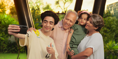 Indian happy old mom dad cute kid standing teen boy hold mobile look phone cam take funny face selfie enjoy home park aged older male elder female lady make video baby girl child fun spend time garden
