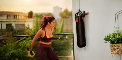 Indian Asian Hindu angry young woman lady do punching on bag outdoor home athletic boxer adult gen z female standing boxing gloves learn sport warm up self fight martial arts class day house - Powered by Adobe