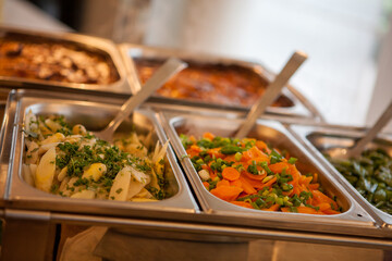 The image is a close up view of a catering buffet, focusing on stainless steel serving trays filled with an assortment of side dishes. In the foreground, there's a tray of boiled potatoes garnished