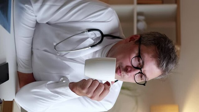 Vertical video. Doctor relaxing. Coffee break. Enjoying moment. Dreamy smiling medical worker man drinking hot beverage from cup resting at hospital workplace.