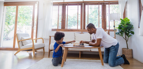 Portrait side view African black father little boy playing chess together, sitting at table, dad and son together educational activity, board logic game at home. Hobby free time, leisure time activity