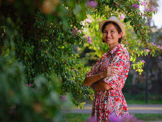 young asian woman stands in sunny green park, admiring blooming bougainvillea bush. The flower's...