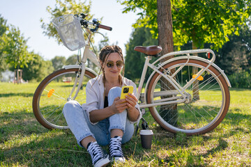 Smiling young hipster woman with bicycle using mobile phone in city park. Talking by video call,...