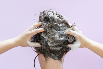 A woman washes her hair with shampoo on lilac background, rear view.
