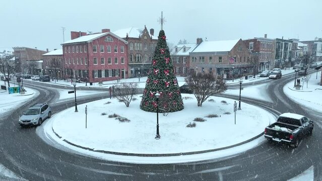 Snowy town square with decorated Christmas tree in traffic roundabout, surrounded by historic buildings. Cinematic aerial shot during December snow storm.