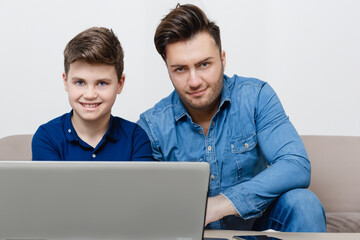 dad and son. young dad sitting with his son on a gray sofa behind a large laptop, dad teaching his son technology, technology concept