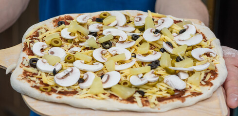 Pizzerman trzyma w dłoniach deskę na której leży pizza z pieczarkami, ananasem i oliwkami gotowa do włożenia do pieca - obrazy, fototapety, plakaty