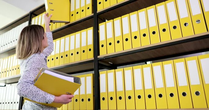 Woman secretary taking out yellow folder from top shelf of bookcase in office 4k movie slow motion . Bureaucracy in business concept