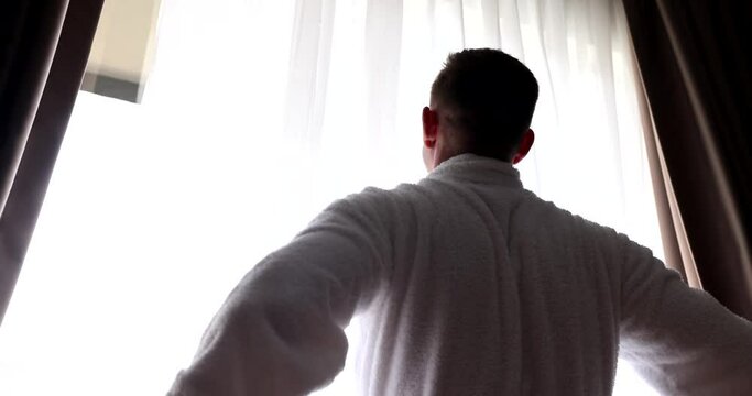 Young man in bathrobe opens curtains at dawn. Rear view of a man with open curtains meets new sunny day in cozy home or hotel