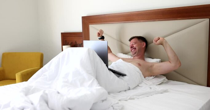 Happy handsome man lies on bed and laughs and reads on message in computer. Good news in morning online