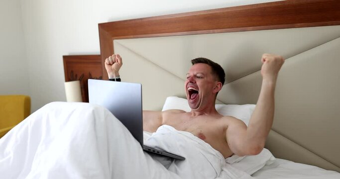 Adult man celebrating success on laptop while lying in bed. Emotions of success of good news and achievements in online network