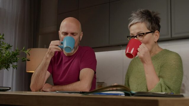 Positive Handsome Mature Man And Charming Middle Aged Woman In Eyeglasses Drinking Coffee, Friendly Talking And Discussing Plan And Design For Home Renovation While Couple Relaxing In Domestic Kitchen