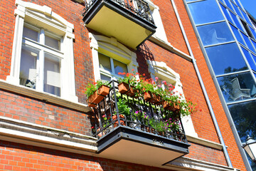 Fototapeta na wymiar Ancient balcony with wrought iron elements and flowers, high windows with white frame and red brick wall. Old street, ancient architecture. Torun, Poland, August 2023