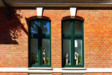 Facade of the historic building with two long windows, green dark frames,  the figures of cats outside the window and a red brick wall. Ancient architecture, old street. Torun, Poland, August 2023 
