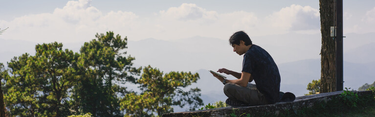 landscape and travel concept with solo freelancer man sit on wooden and use tablet work from outdoor with layer of mountain background
